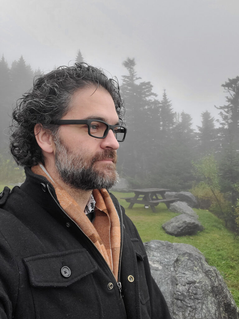 Image of Logan Kendell (aka Vincent Blackwood), a man with a graying beard wearing black glasses and a black peacoat and scarf, standing in the mist at the top of Stowe Mountain in Vermont.