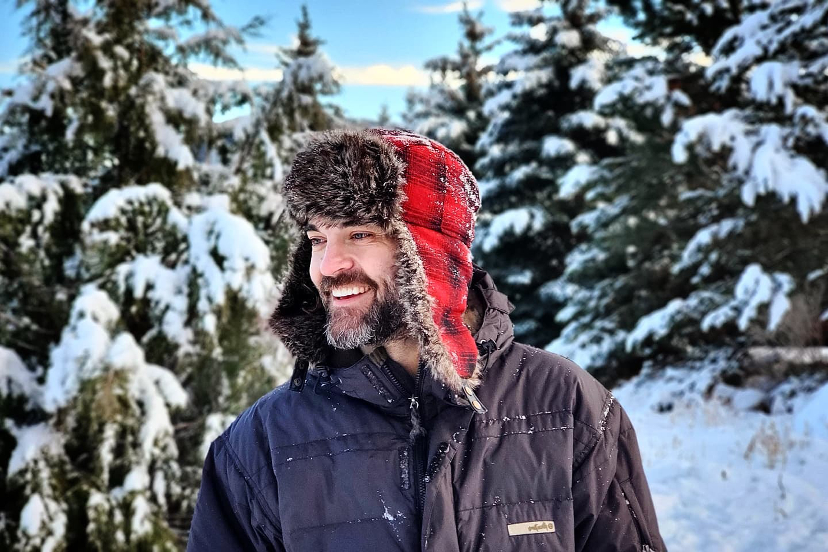 Image of Logan Kendell (aka Vincent Blackwood) standing in a snowy pine forest wearing a fur trimmed lumberjack style winter hat and down ski coat.